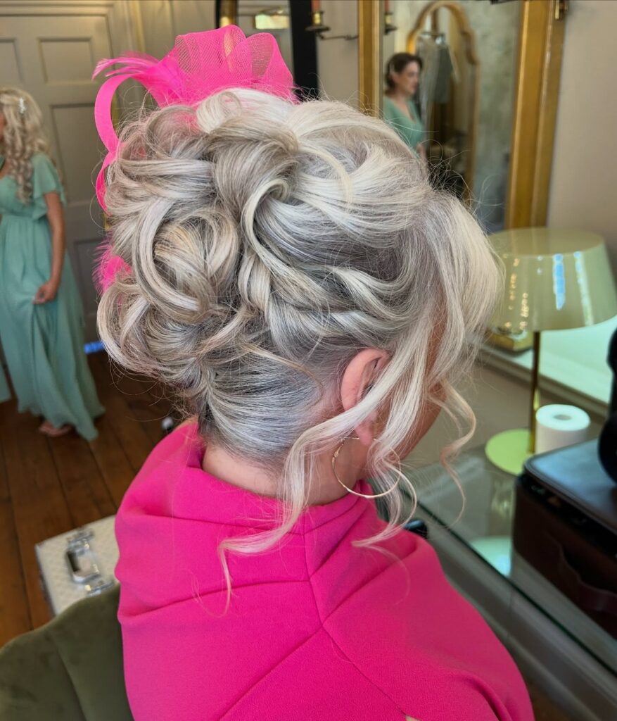 A woman with styled, curly silver hair in an updo is seen from the back, adorned with a large hot pink fascinator.