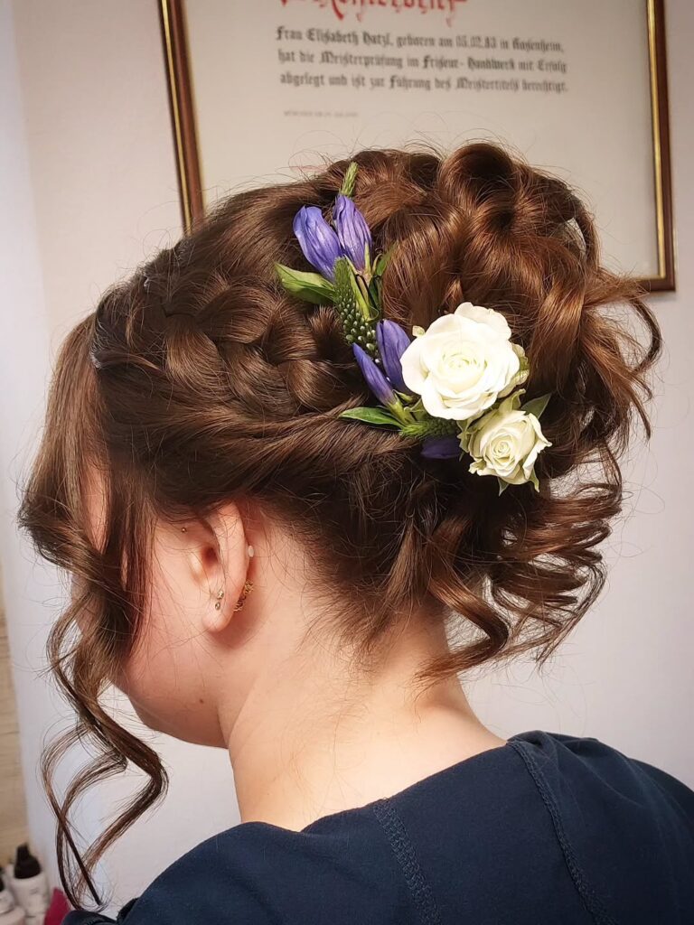A woman with dark brown hair in a braided updo with cascading curls decorated with white roses and purple flowers, shown from the side back.