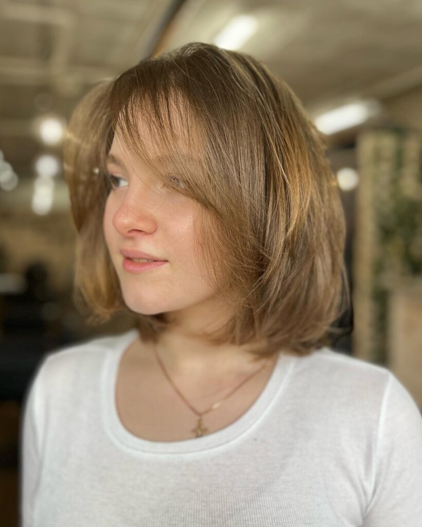 A woman with a golden bronde chin length bob and wispy bangs looks off to the side.