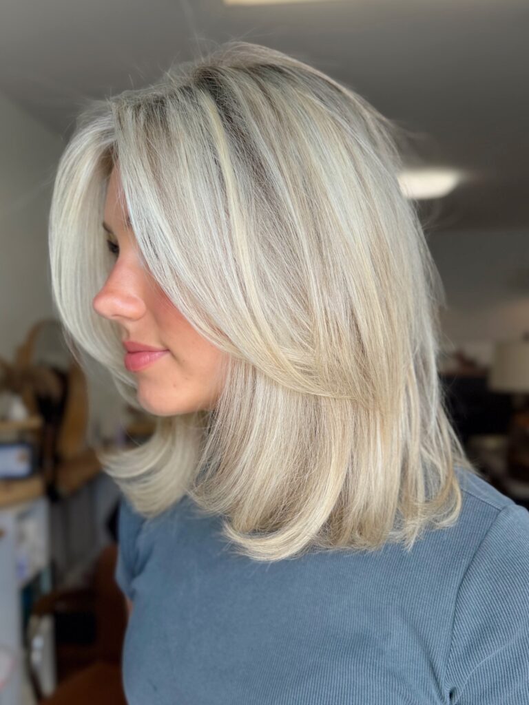 A woman with an icy platinum blonde layered lob and dark root melt is shown in profile.
