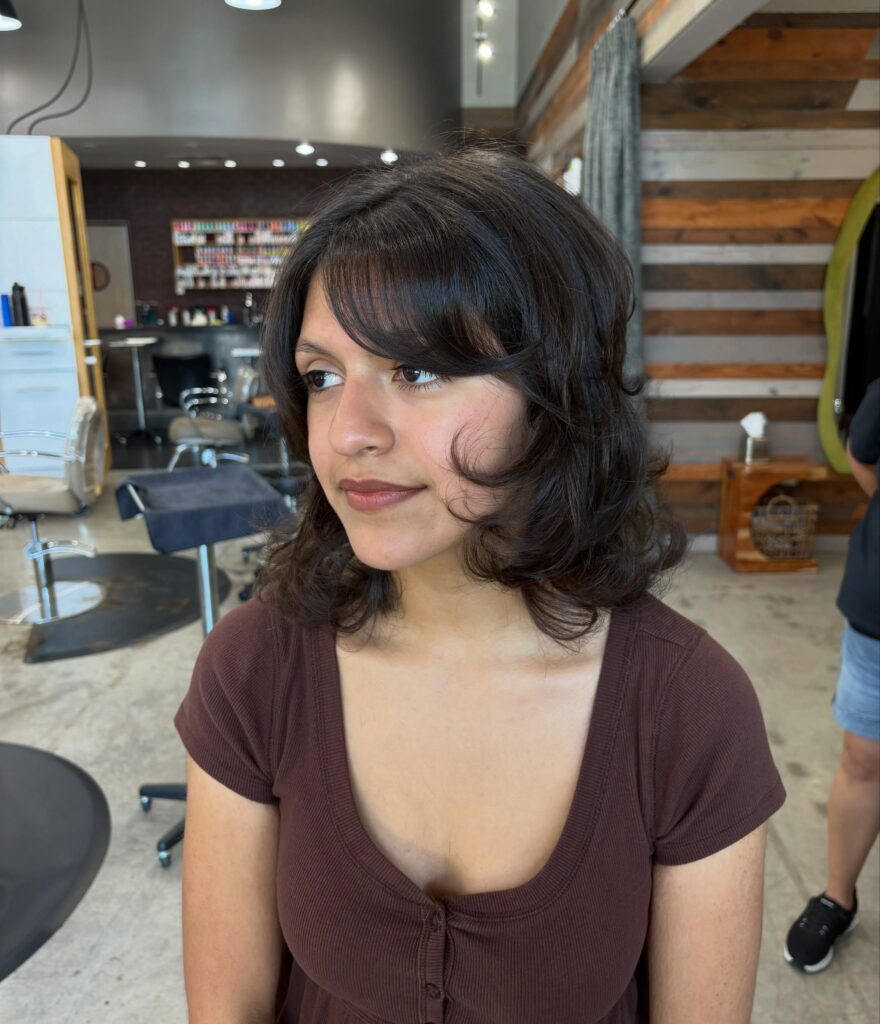 A woman with dark brunette wavy hair and side swept bangs smiles gently in a salon.
