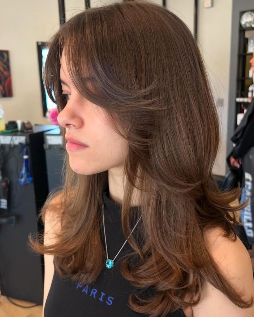 A woman with long chestnut brown layered hair featuring flipped ends and curtain bangs is shown in profile.