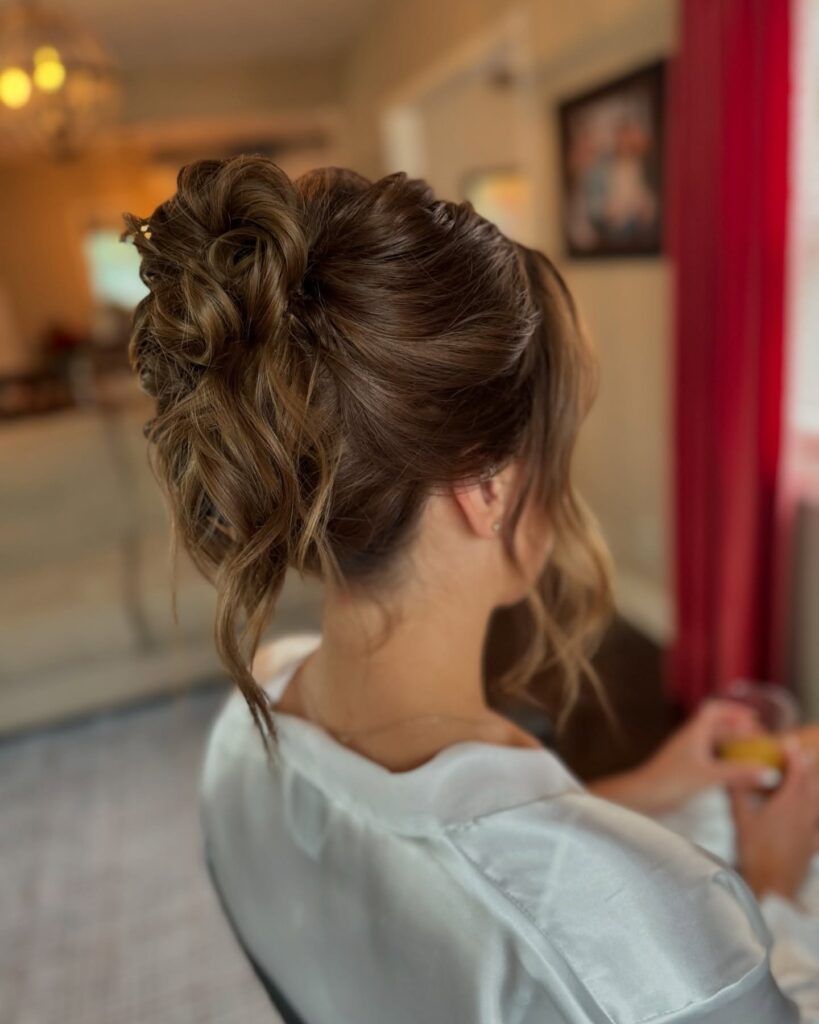 A woman with thick light-brown hair styled in a high wavy updo with dimensional highlights and face framing tendrils.