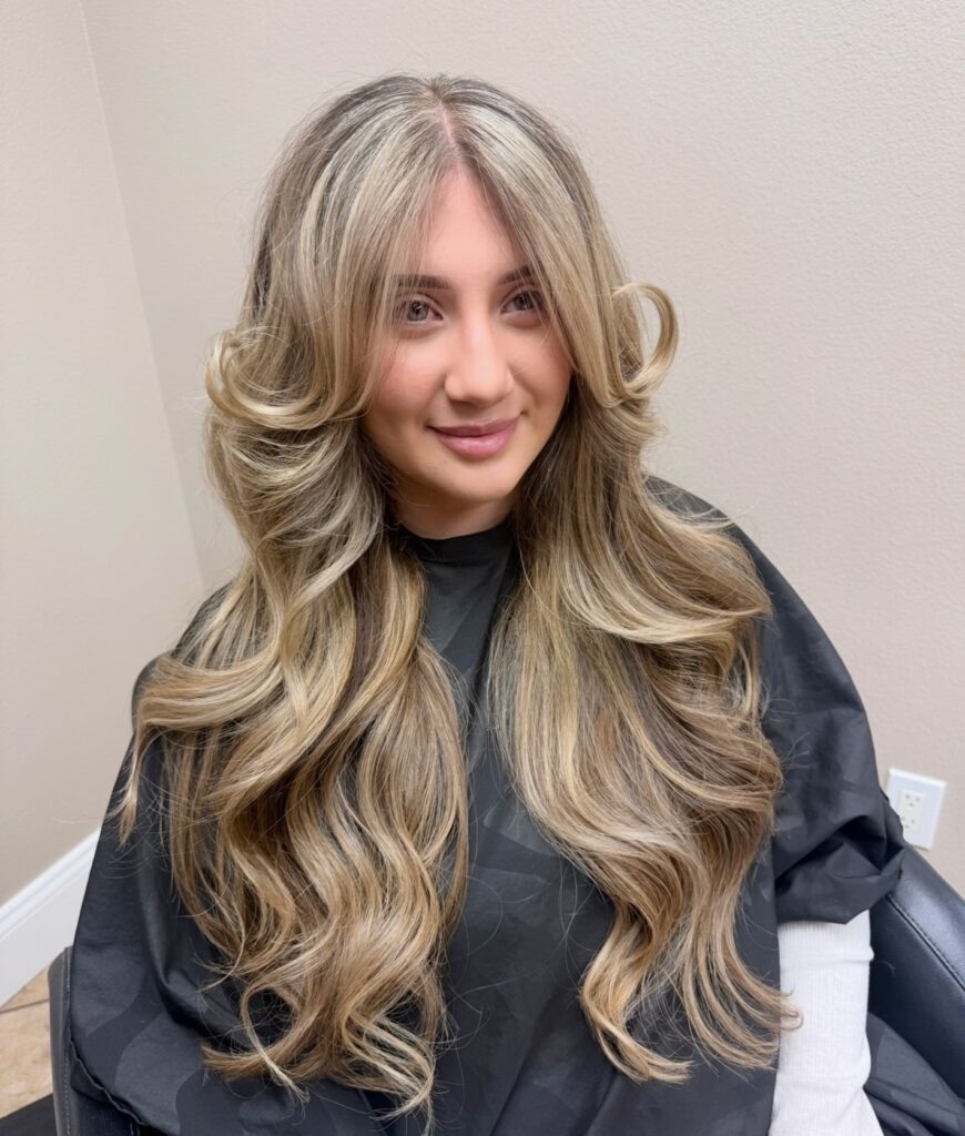 A woman with extra long golden blonde curly layered hair and curled face framing pieces.