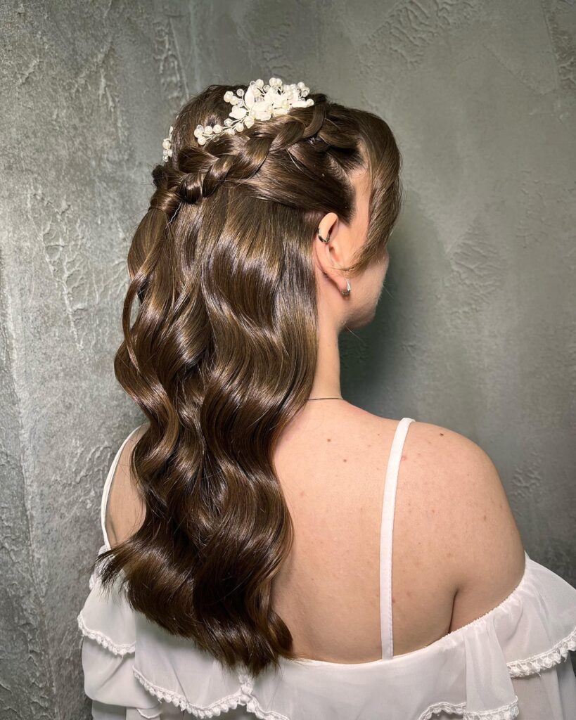 Back view of a woman with long glossy chestnut brown waves styled in a half up crown braid decorated with pearl and crystal hair pins.