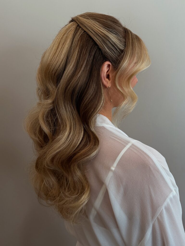 Side view of a woman with long golden blonde voluminous waves styled in a half up twist at the crown with a curled face framing strand.