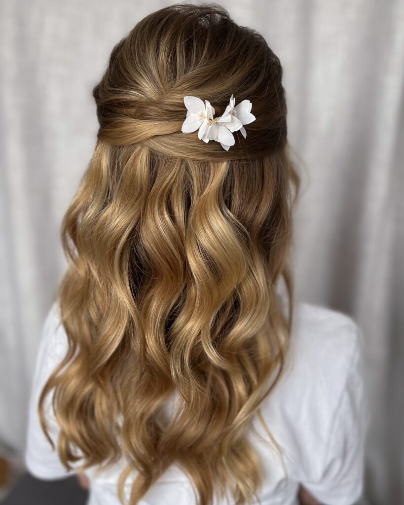 Back view of a woman with long golden blonde wavy hair styled in a half up twist at the crown with a small white flower hair accessory.