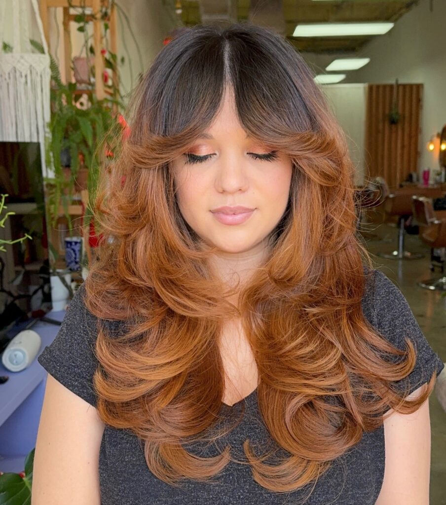 Woman with long layered hair in a dark brown to copper ombre, center parted curtain bangs and bouncy voluminous curls with flipped out ends.