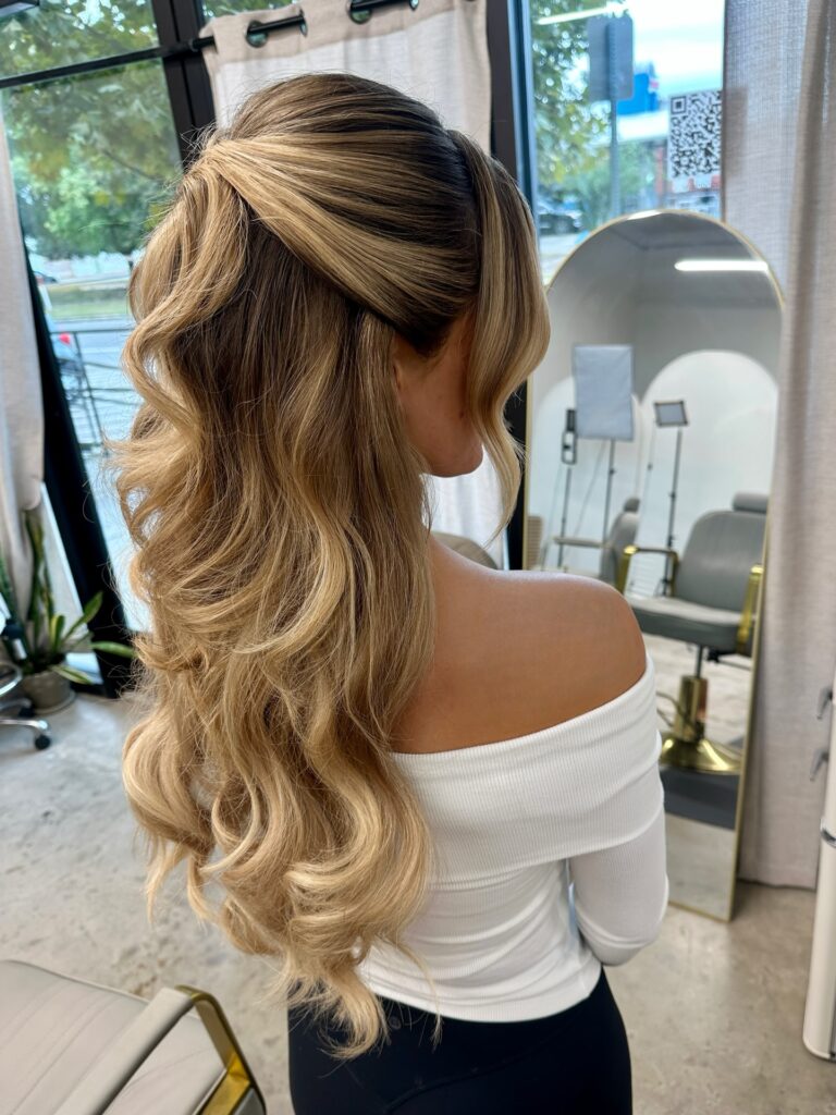 Back view of a woman with long blonde hair styled in a voluminous half up style with a lifted crown and big soft waves.