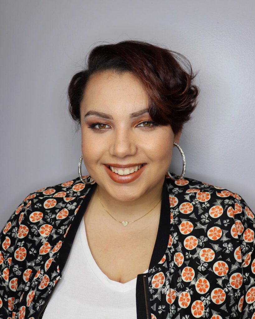 A smiling woman with a mahogany-colored pixie cut styled with voluminous curls on top and a deep side part.
