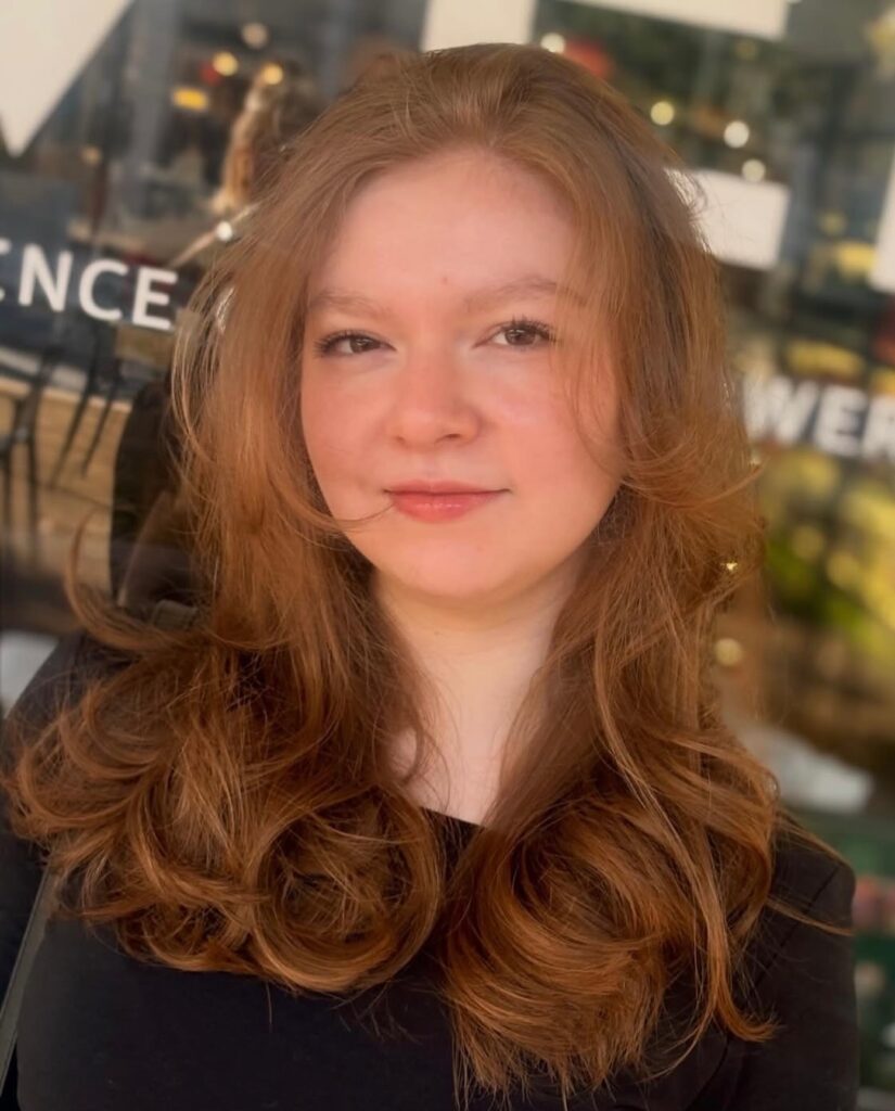 A woman with long, voluminous copper hair featuring face-framing layers and bouncy, curled ends.