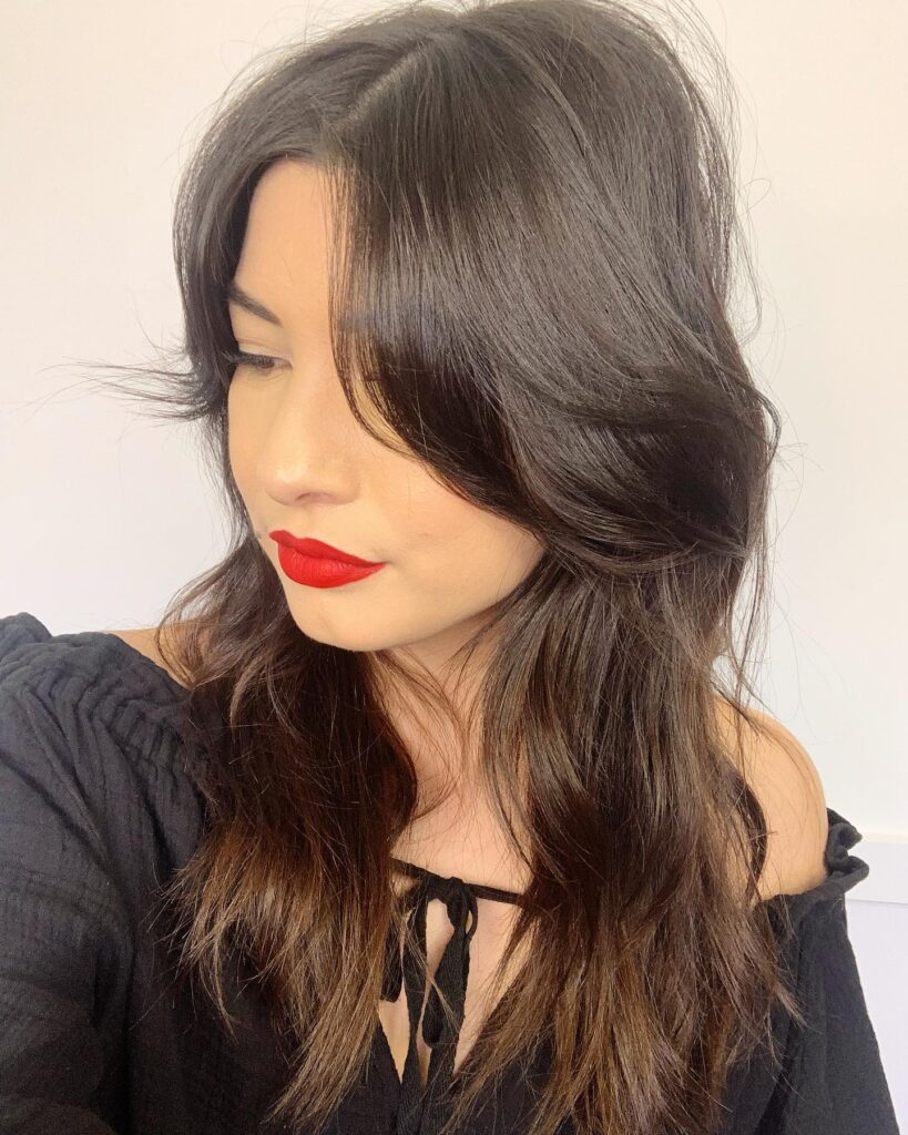 A woman with long, wavy, dark brown hair and face-framing curtain bangs wearing red lipstick.