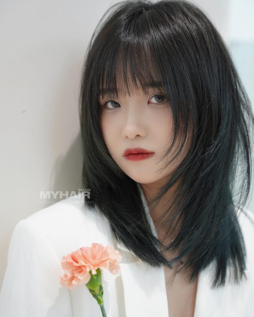 A woman with a shaggy black wolf cut and wispy fringe bangs holding a pink flower.