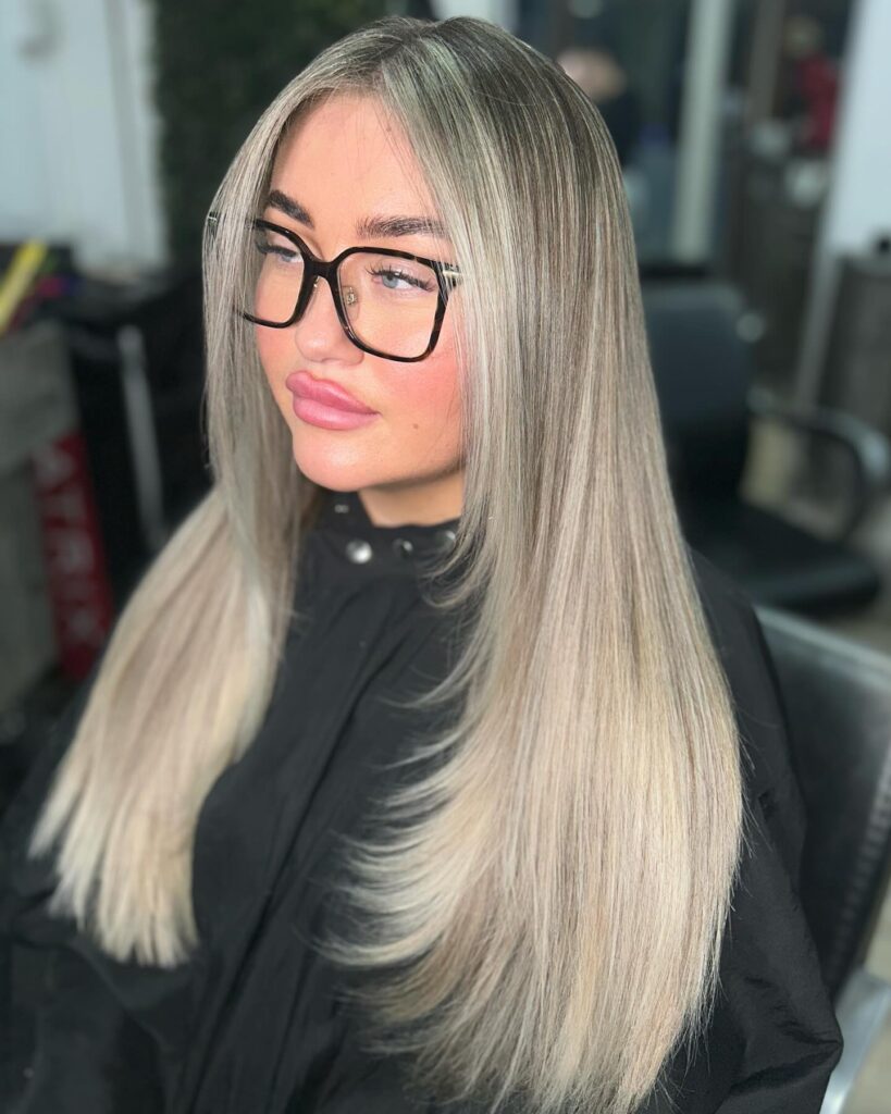 A woman with long, straight ash-blonde hair featuring face-framing layers and soft curtain bangs.