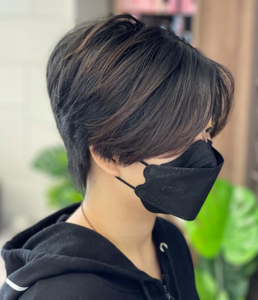 A person with a dark brown tapered pixie cut featuring long side-swept bangs and voluminous layers on top.