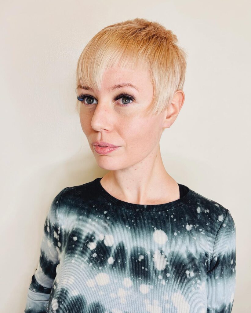 A woman with a very short strawberry-blonde pixie cut featuring a micro fringe and textured layers on top.