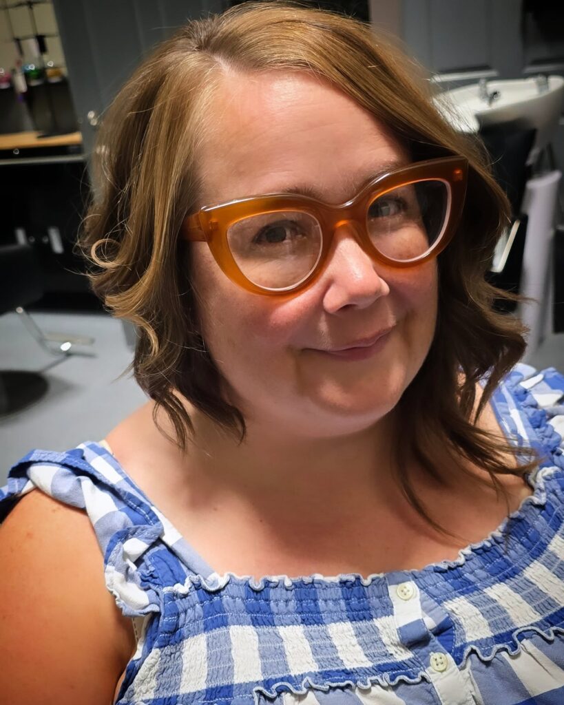 A smiling woman with a shoulder-length honey blonde lob featuring soft waves and a side-swept front while wearing orange-rimmed glasses.