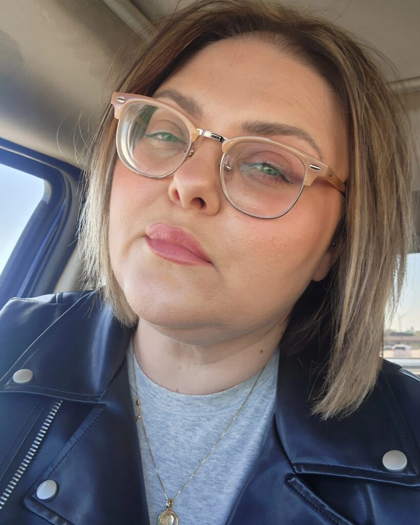 A woman with a straight ash-brown lob and a deep side part, wearing glasses and a leather jacket.