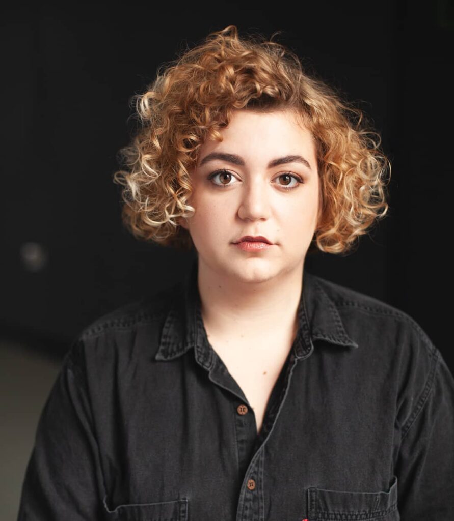 A woman with a voluminous chin-length curly bob in golden blonde tones with a deep side part.