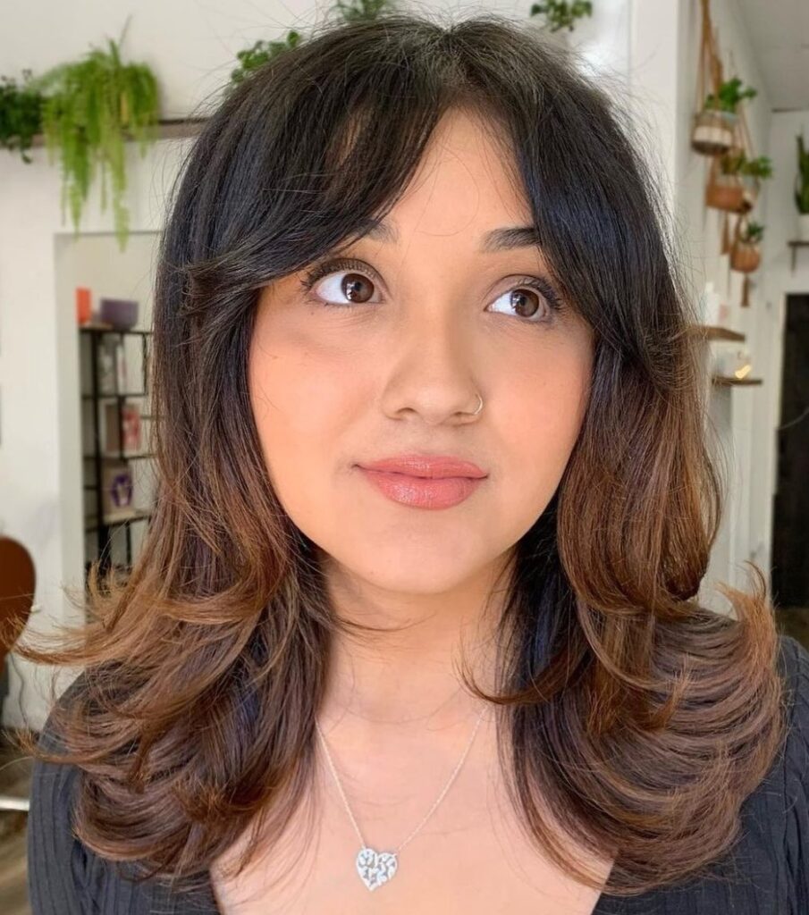A woman with medium-length brown hair featuring a caramel ombre, voluminous flipped layers, and curtain bangs.