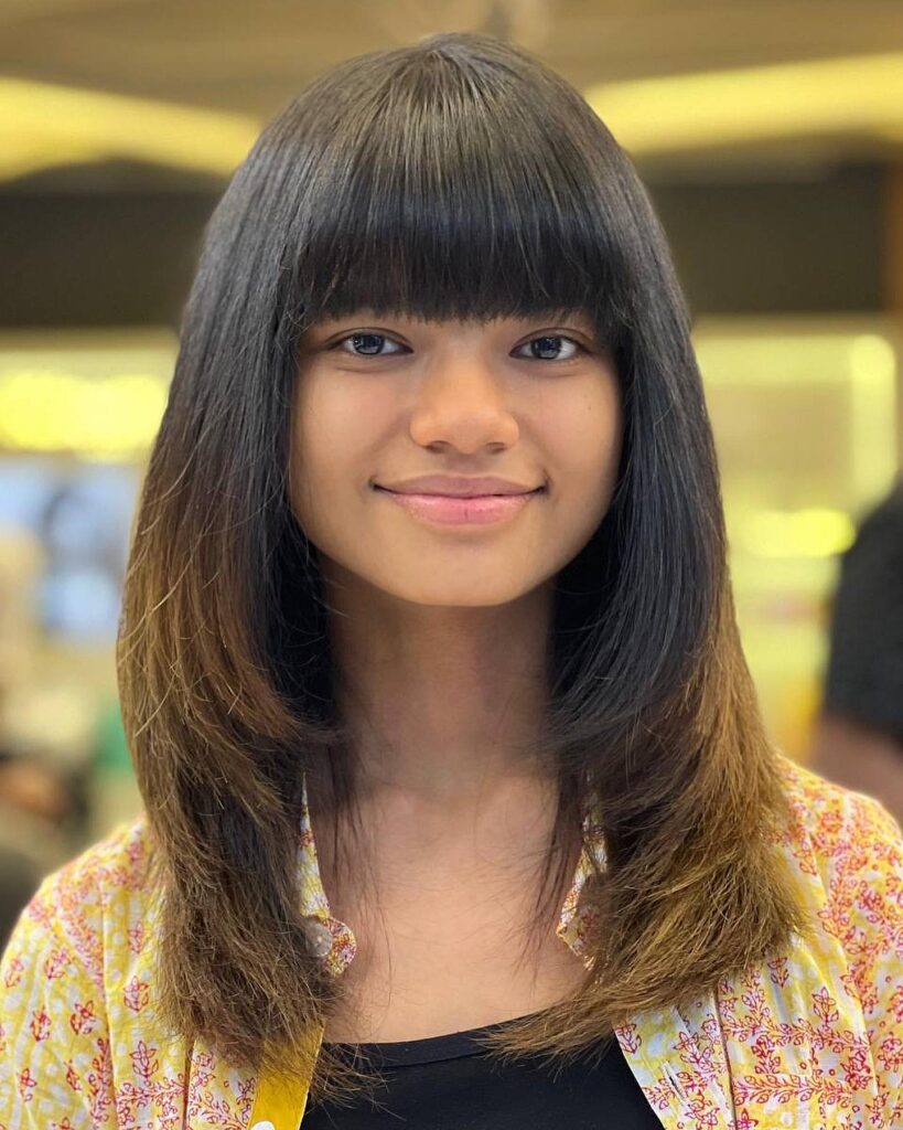 A woman with mid-length dark hair featuring a heavy straight-across fringe and soft, rounded layers.