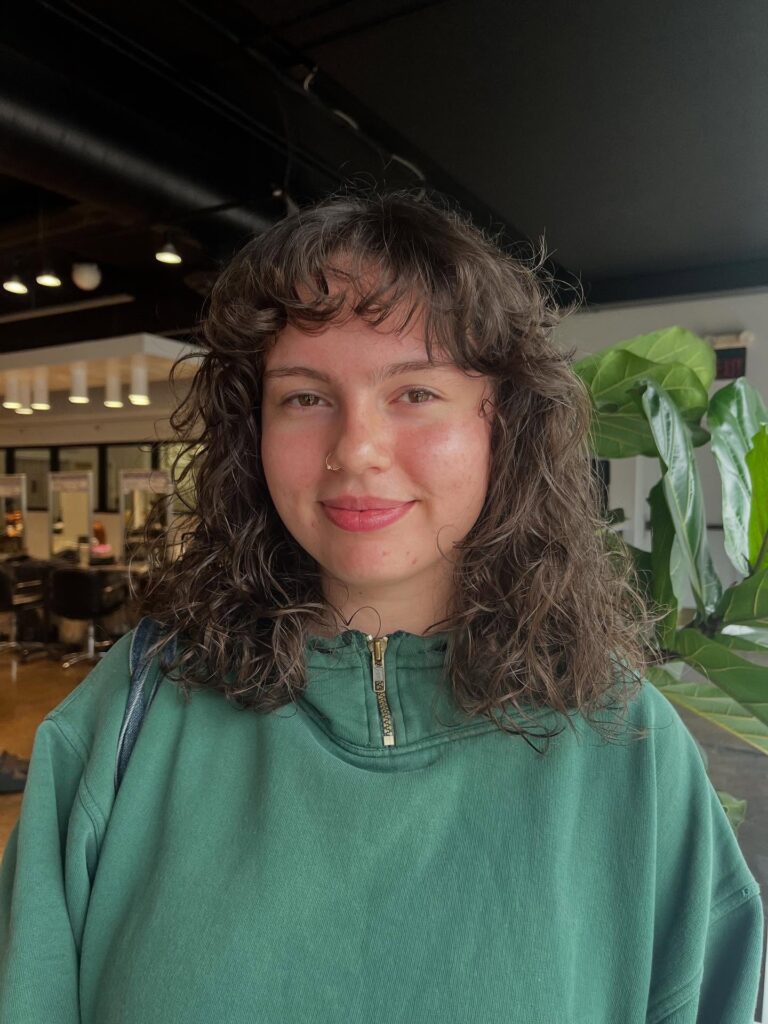A woman with shoulder-length curly brunette hair featuring textured layers and a curly fringe.