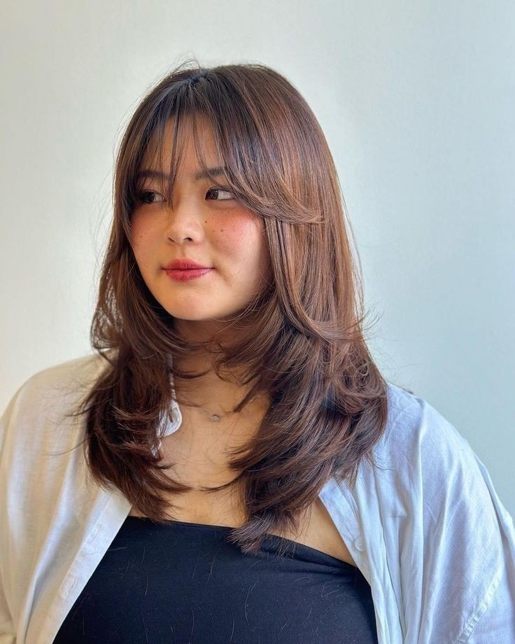 A woman with medium-length chocolate-brown hair styled in voluminous butterfly layers with wispy bangs.