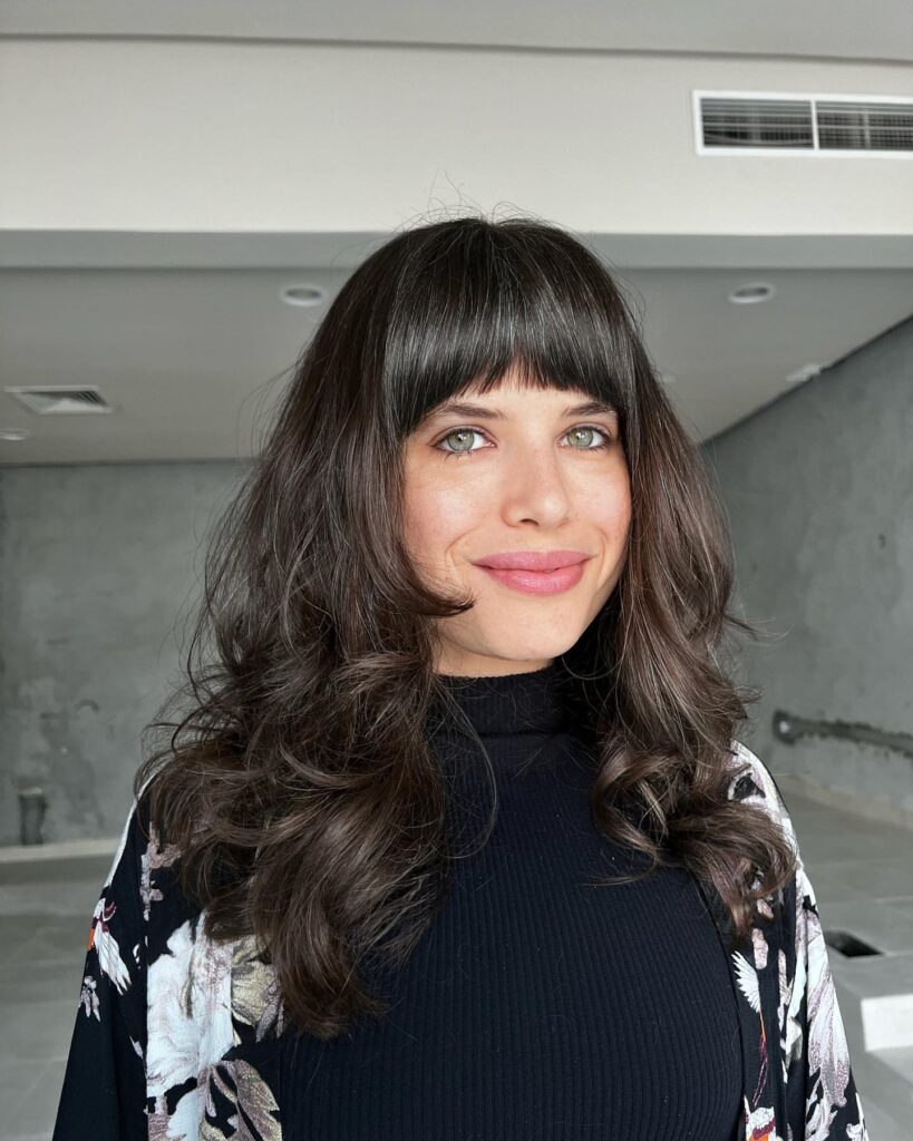 Layered dark brown haircut with voluminous waves and a full fringe.