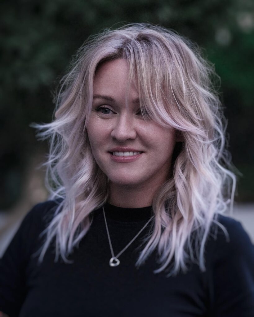 A woman smiling with shoulder-length icy blonde hair styled in beachy waves and curtain bangs.