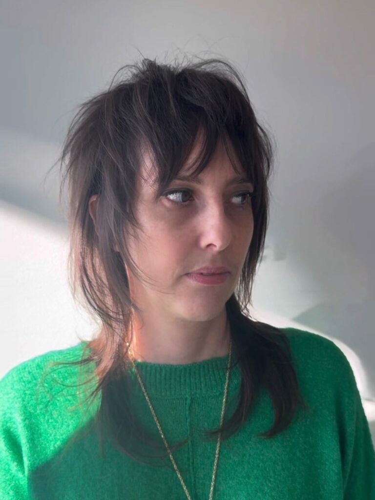 A woman wearing a green sweater with a dark brunette wolf cut featuring shaggy layers and choppy bangs.