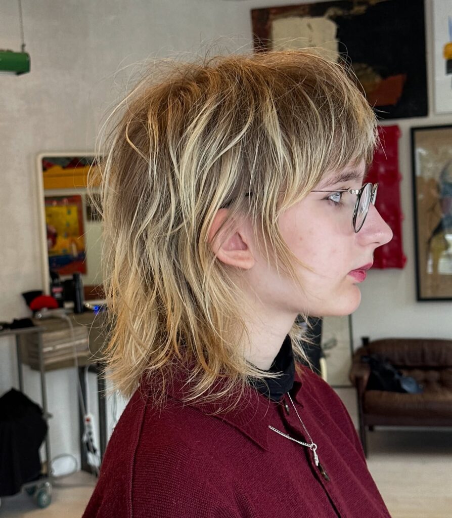 A woman with a textured blonde wolf cut featuring heavy shaggy layers and full fringe bangs.
