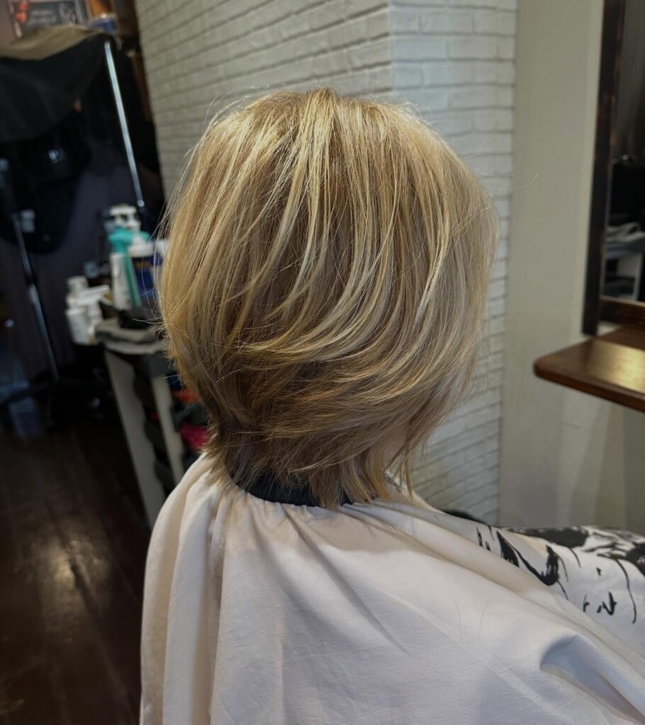 A back view of a woman with a short graduated blonde bob featuring honey highlights and piecey stacked layers.