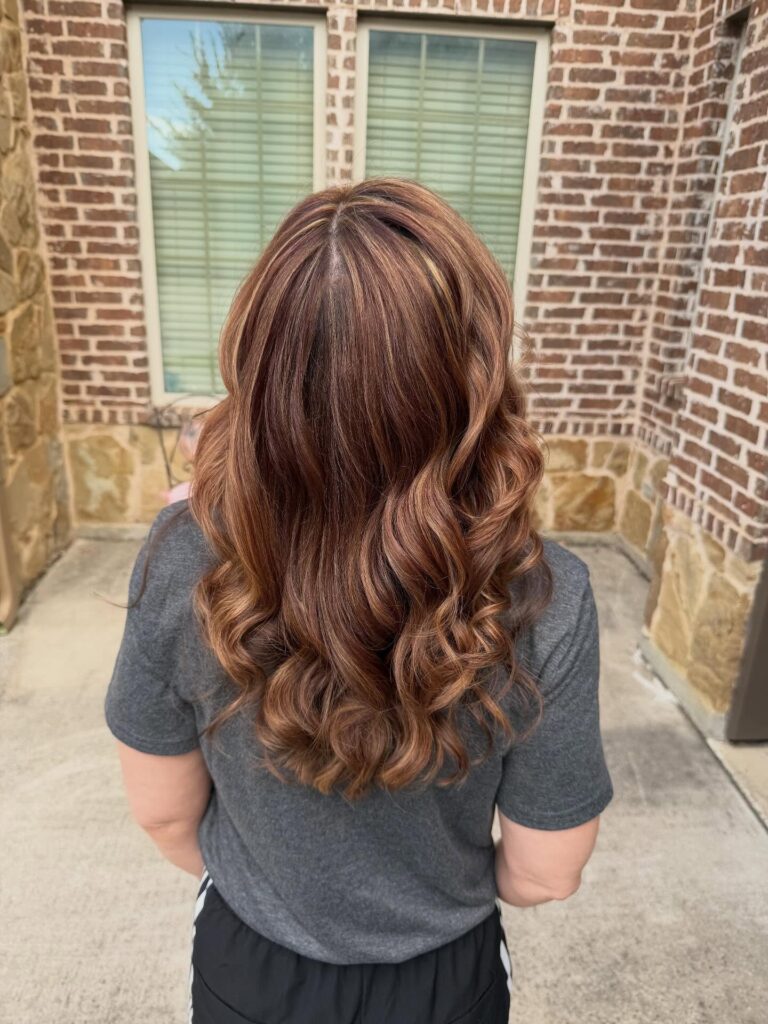 The back view of a woman with long chestnut hair styled in large bouncy curls with golden highlights.