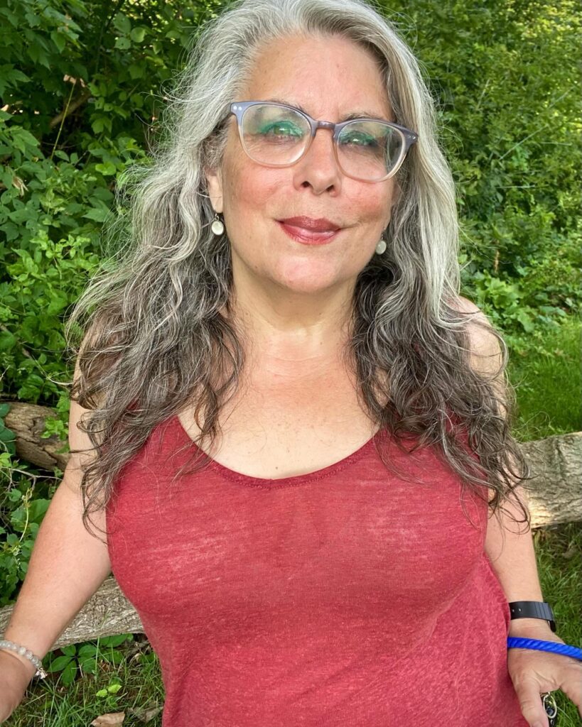 A woman with long salt-and-pepper hair styled in natural loose waves with bright silver highlights around the face.