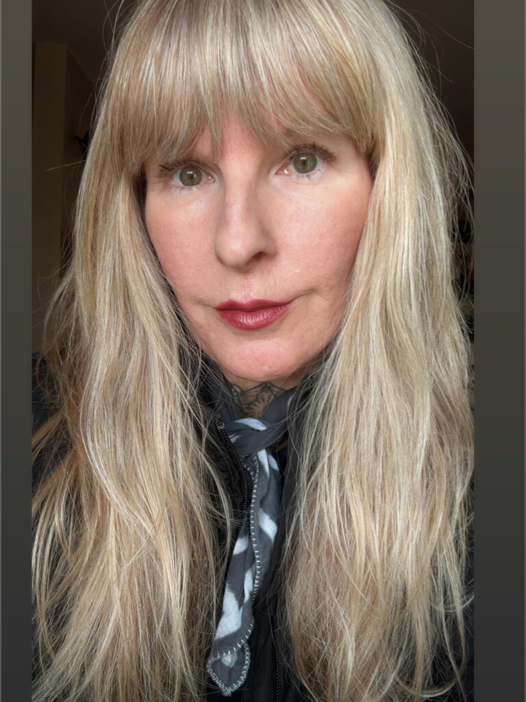 A woman with long ash-blonde hair featuring a full wispy fringe and textured layers.
