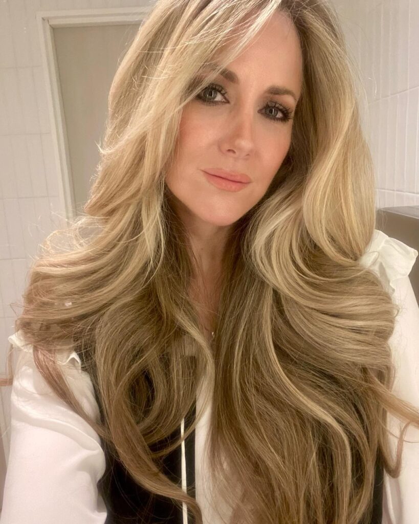 A woman with long, voluminous champagne-blonde hair styled in thick, glamorous waves with face-framing layers.
