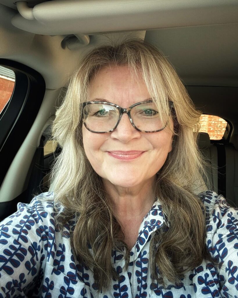 A smiling woman with long ash brown hair featuring soft blonde highlights and face-framing curtain bangs.