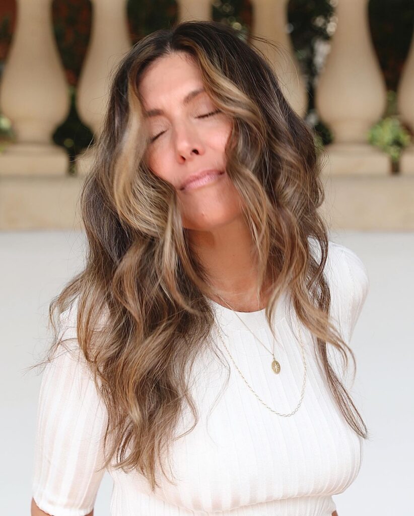 A woman with long, wavy ash-brown hair featuring bright blonde money piece highlights and soft, textured layers.
