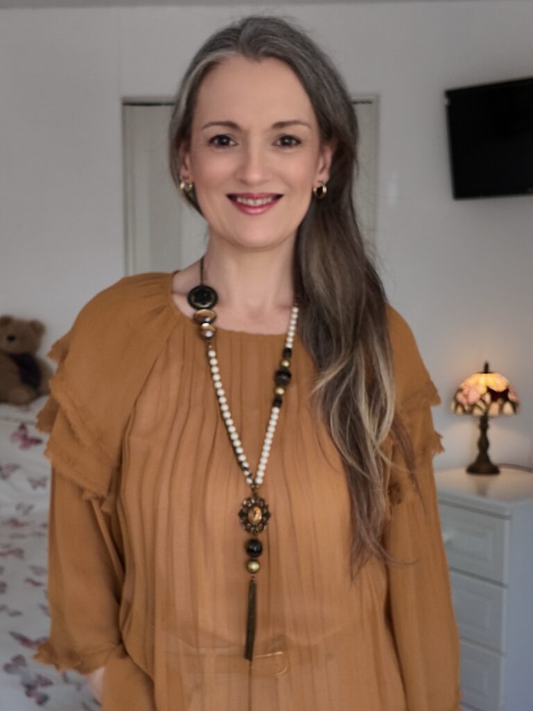 A smiling woman with long, straight pewter hair swept over one shoulder, wearing a mustard yellow blouse.