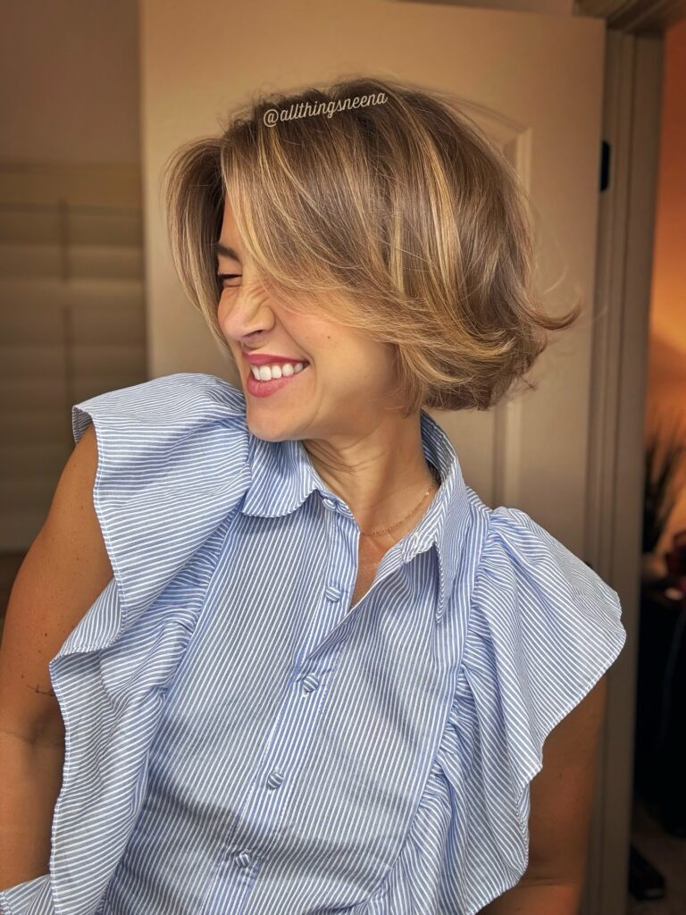 A smiling woman wearing a blue striped shirt with a voluminous honey-blonde bob that features flipped-out ends and sweeping layers.