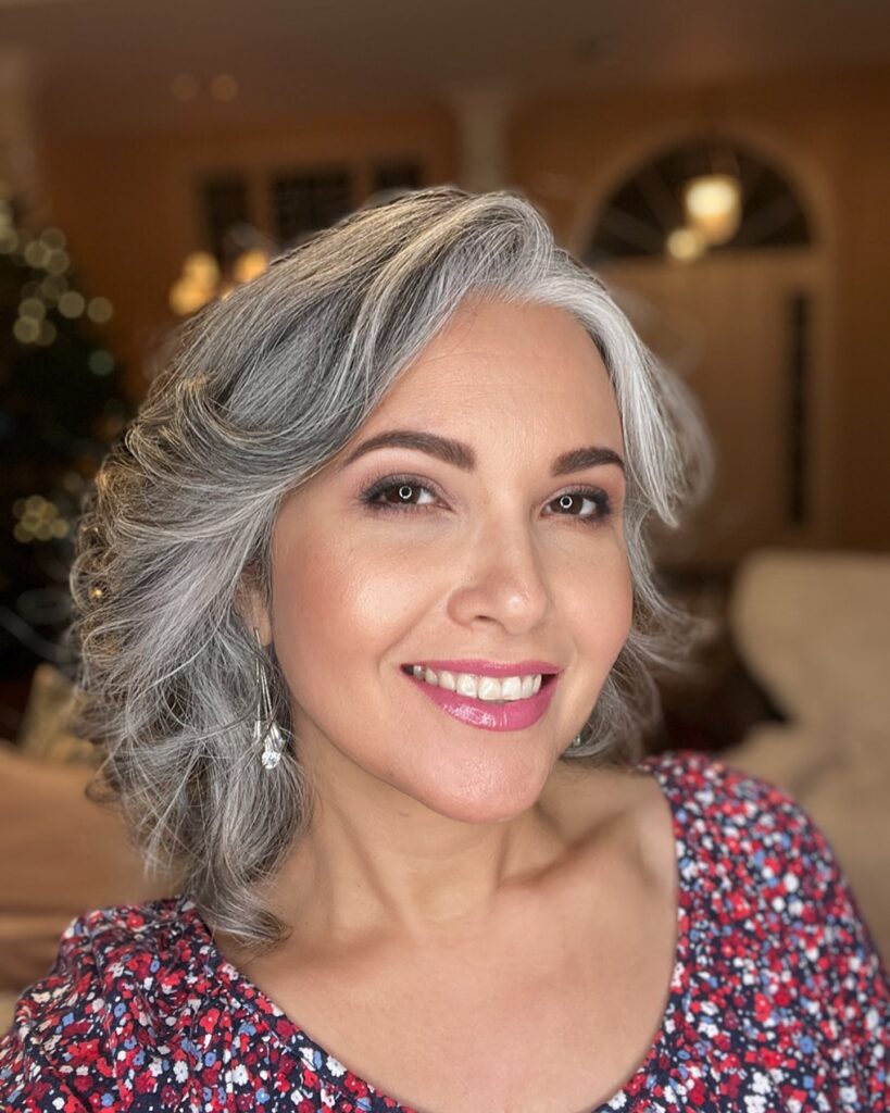 A smiling woman with a voluminous shoulder-length silver hairstyle featuring soft layered waves and a side part.