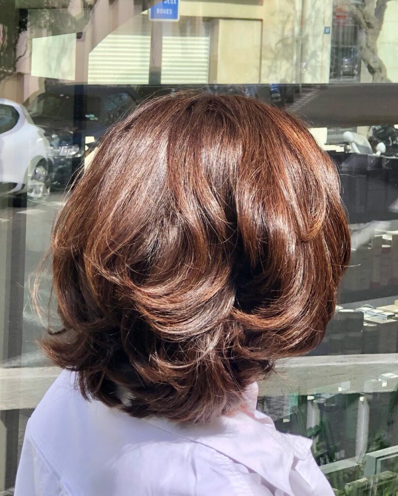 A back view of a woman with a voluminous neck-length chestnut brown bob featuring multiple feathered layers and high shine.