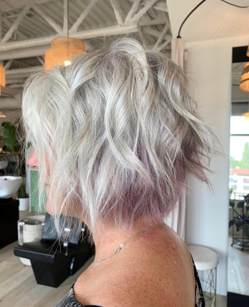 A side view of a woman with a messy, textured silver bob featuring lavender purple peekaboo highlights underneath.