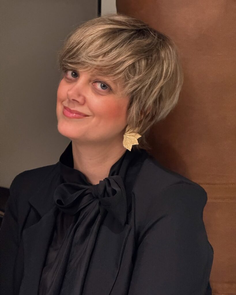 A woman with a honey-blonde shaggy crop hairstyle featuring thick side-swept bangs and textured layers.