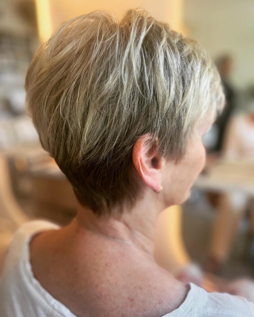 A side profile of a woman over 50 with a tapered ash blonde pixie cut featuring short layers at the nape and volume on top.