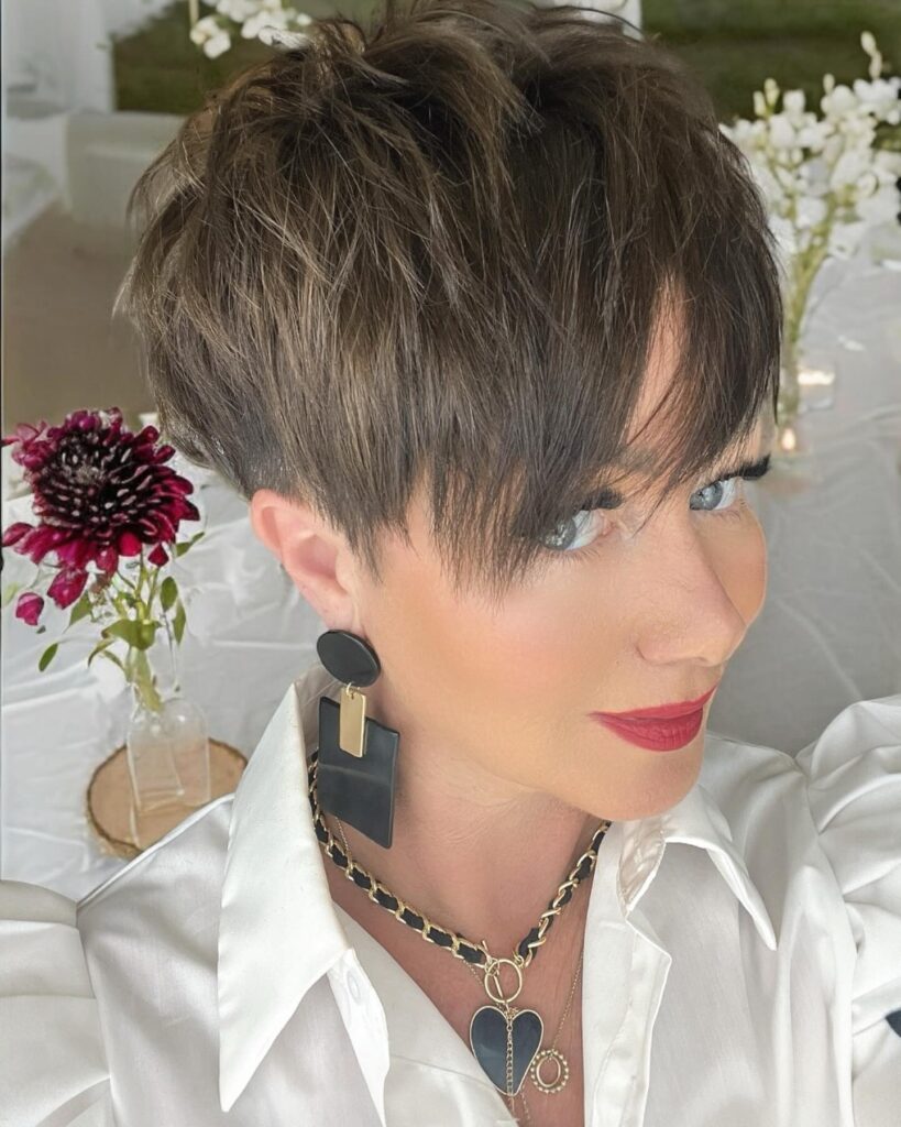 A woman with a short, edgy brunette pixie cut featuring an undercut and long choppy bangs.