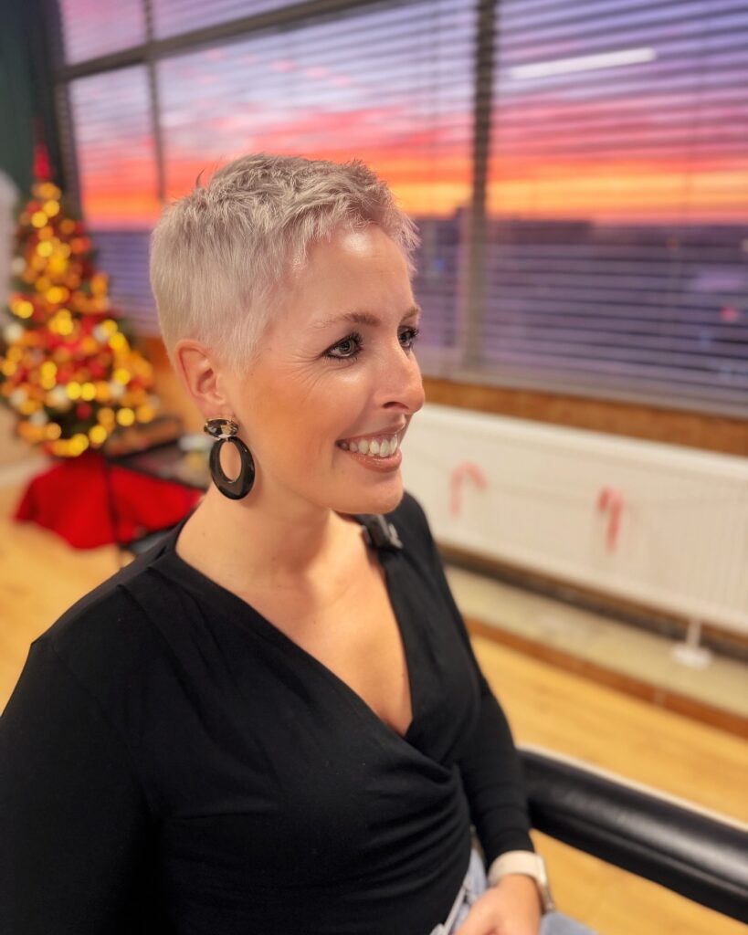 A woman over 50 with an ultra-short platinum blonde buzz cut smiling in a side profile.