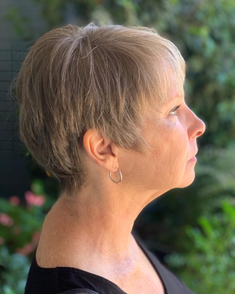 A side profile of a woman over 50 with a soft mushroom brown pixie cut featuring feathered layers and a tapered nape.