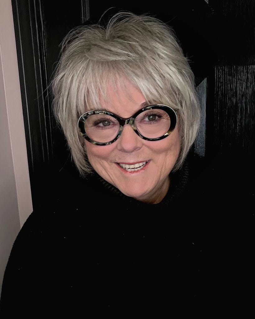 A short layered messy bob with full fringe and light blonde texture, styled with glasses.
