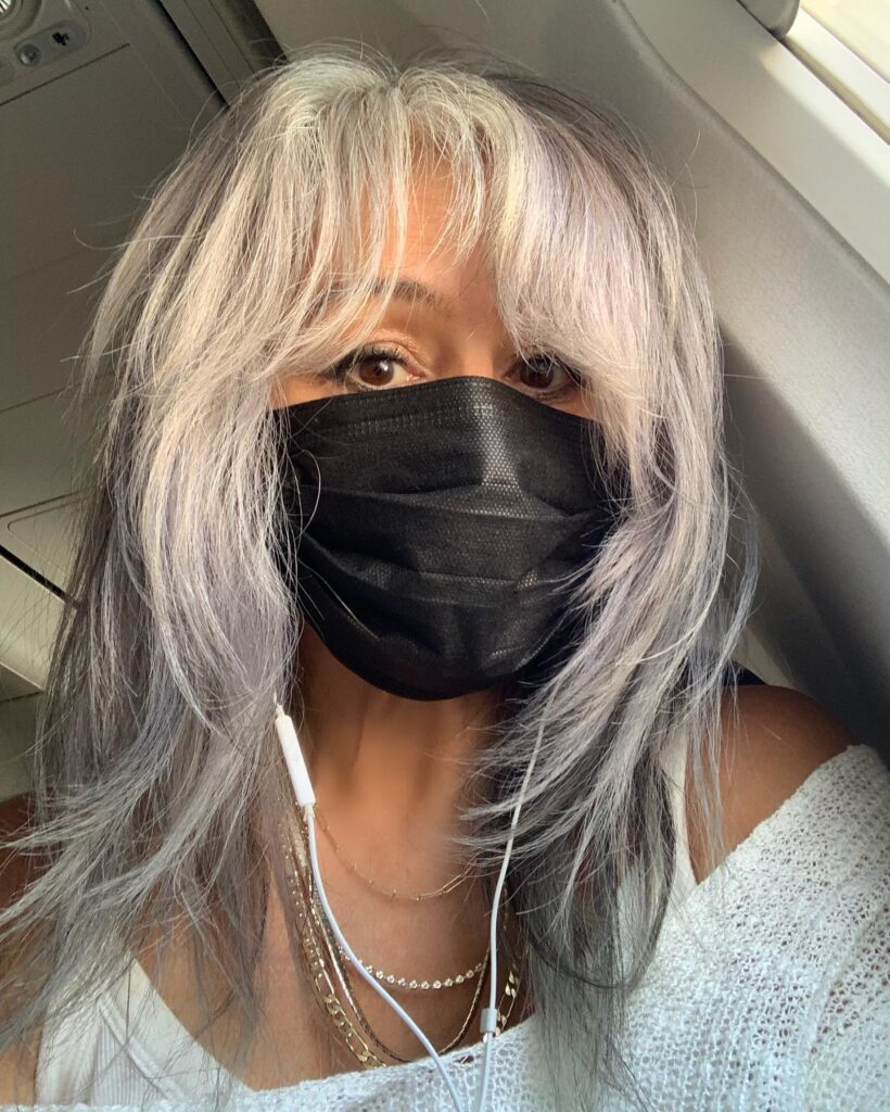 A woman wearing a black mask with long, silver, layered hair featuring shaggy, feathered ends and a soft, wispy fringe.